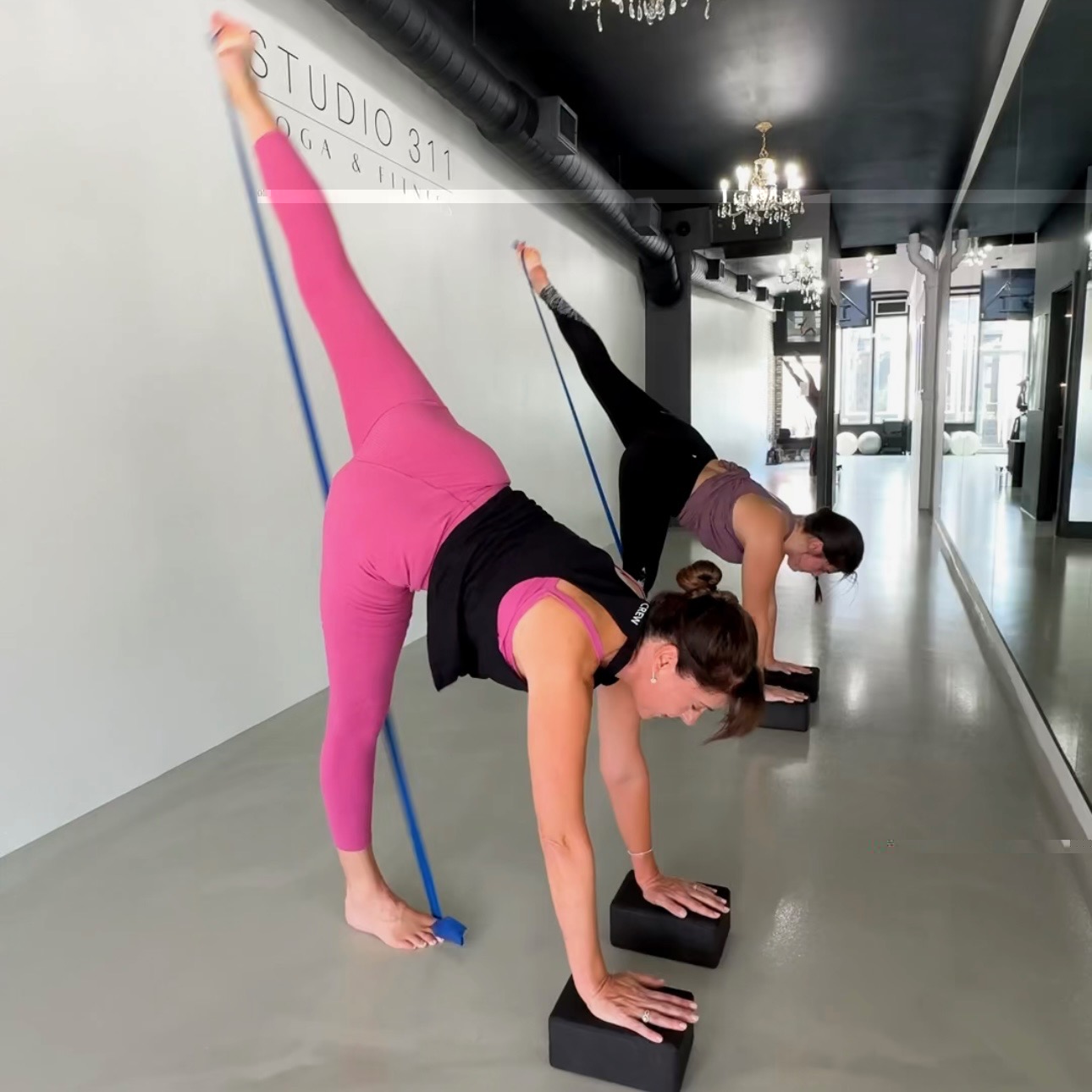 Two women practice flexibility and mobility exercises at Studio 311 Yoga & Fitness. Each uses a resistance band looped under one foot and anchored by their hands on yoga blocks, extending the opposite leg high behind them in a controlled stretch. The bright, modern studio features polished floors, chandeliers, and a mirrored wall that reflects their focused movement