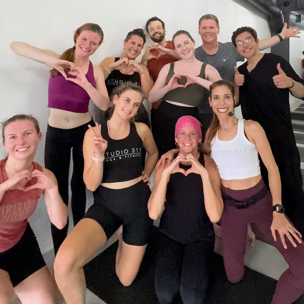 Inside the studio at Studio 311 yoga & Fitness in Daytona Beach, FL a joyful group of nine people in workout attire pose indoors, forming heart shapes with their hands, conveying a sense of community and positivity.