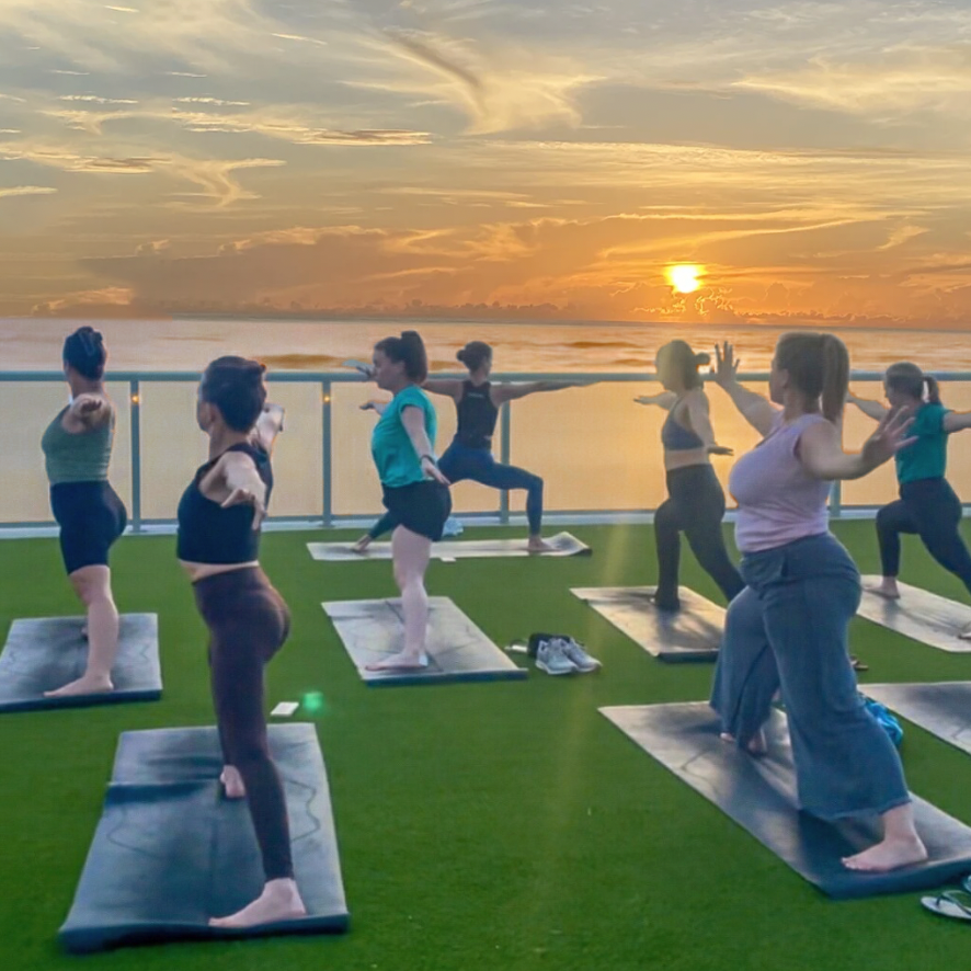 Women practice yoga outdoors on mats at sunrise facing a calm sea. The scene is serene and focused, with a warm, golden sky in the background. Renaissance Hotel Daytona Beach. Led by Sherri Meyers