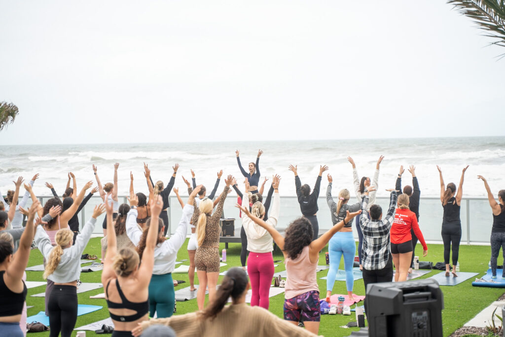 A large group of people from the New Smyrna Beach Social club practice yoga outdoors facing the ocean. They are led by Sherri Meyers of Studio 311 Yoga & Fitness. Their arms are raised under a cloudy sky, conveying a peaceful and energetic atmosphere. This on the event lawn at Renaissance Daytona Beach Oceanfront Hotel.