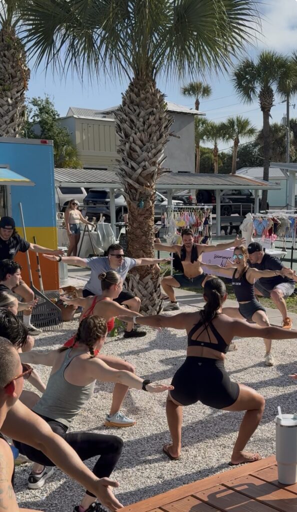 A group of runners from the New Smyrna Run club are doing outdoor yoga under palm trees, surrounded by colorful stalls. The mood is energetic and cheerful. They are led in their pre-run stretch by Sherri Meyers of Studio 311 Yoga & Fitness. 