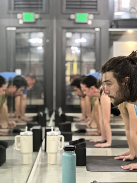 A group of people doing plank poses in a studio, reflected in mirrors. Water bottles and towels are on the floor, creating a focused and calm atmosphere.