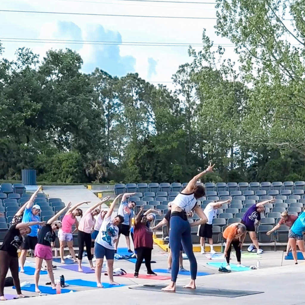 A yoga class outdoors with instructor Sherri Meyers and diverse participants stretching on mats. The setting is a green park with clear skies and a relaxed atmosphere. This is part of Healthy Port Orange.