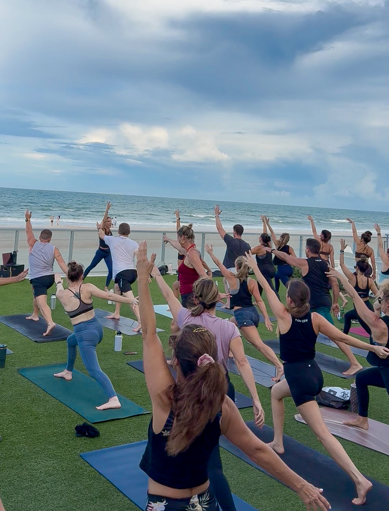 “Inside Flow: Ocean Edition” yoga class on a beachside deck at Renaissance Daytona Beach, FL, with a group stretching on mats beside the sea under a cloudy sky; energetic, communal mood. The Inside Flow yoga class is taught by Sherri Meyers of Studio 311 Yoga.